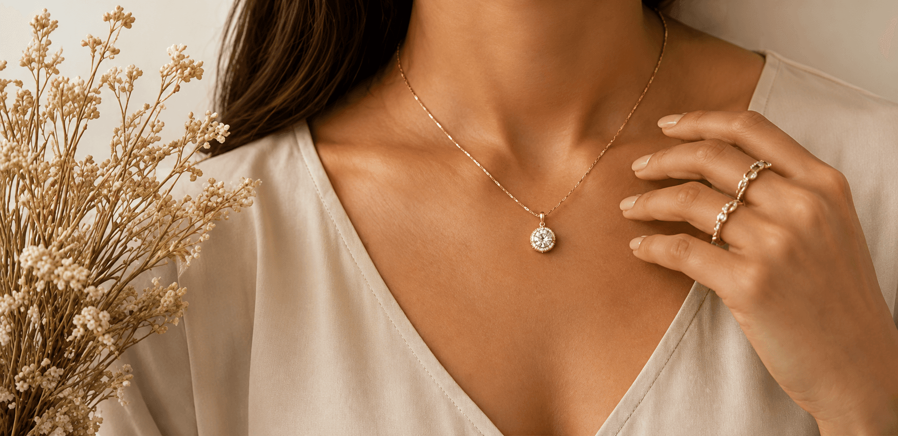 Woman wearing a delicate gold diamond pendant and stacked rings beside dried flowers